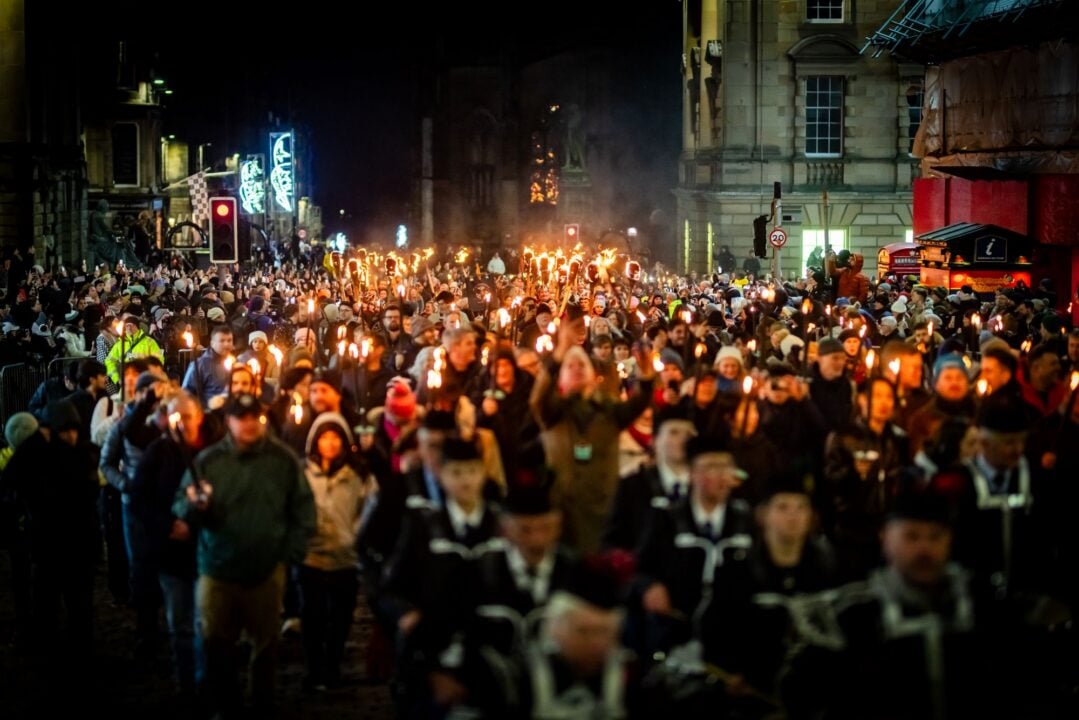 A crowd of people walking through a street at night, some of whom are holding firelit torches.
