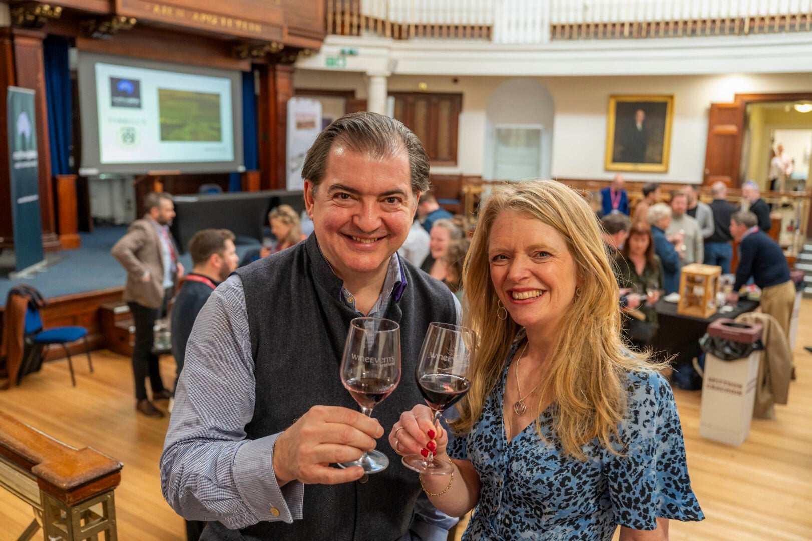 A couple toasting with glasses of red wine. Behind them are a room full of people.