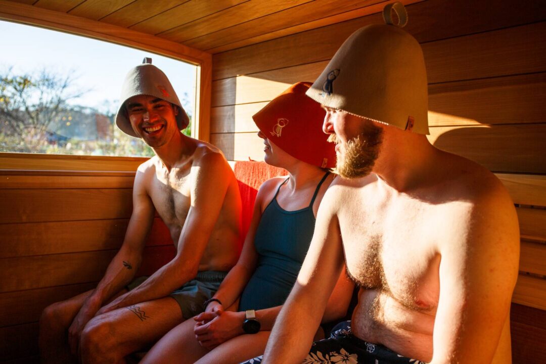 Three people sitting inside a sauna with hats on. Sun is shining through the window.