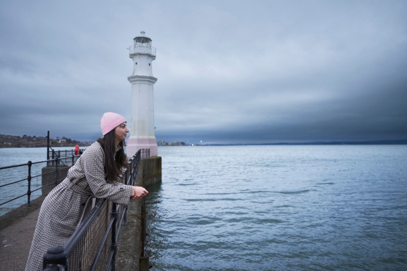 A photo of a woman looking out over Newhaven Harbour with a lighthouse behind her.