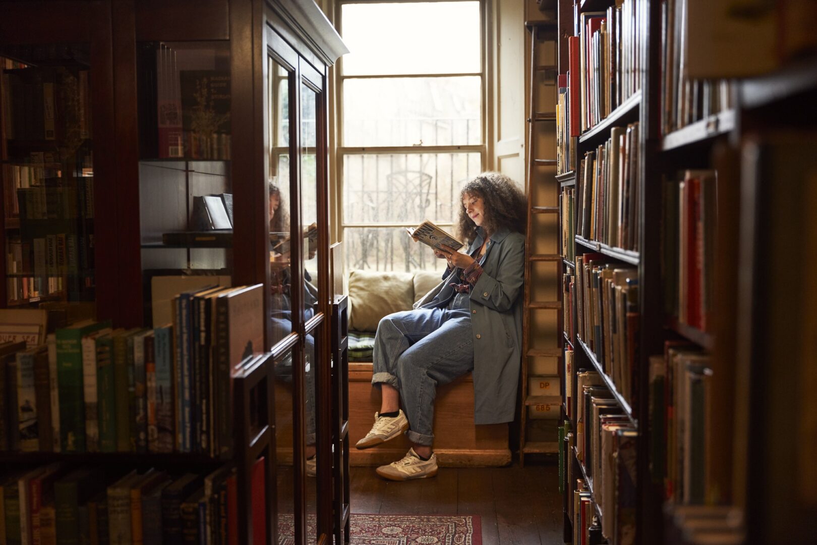 A lady sitting on a window seat in a bookshop reading a book