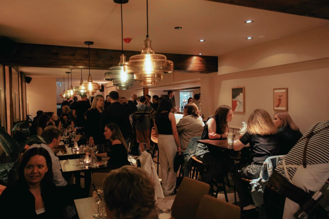 A bar full of people. seated round individual tables, and standing.