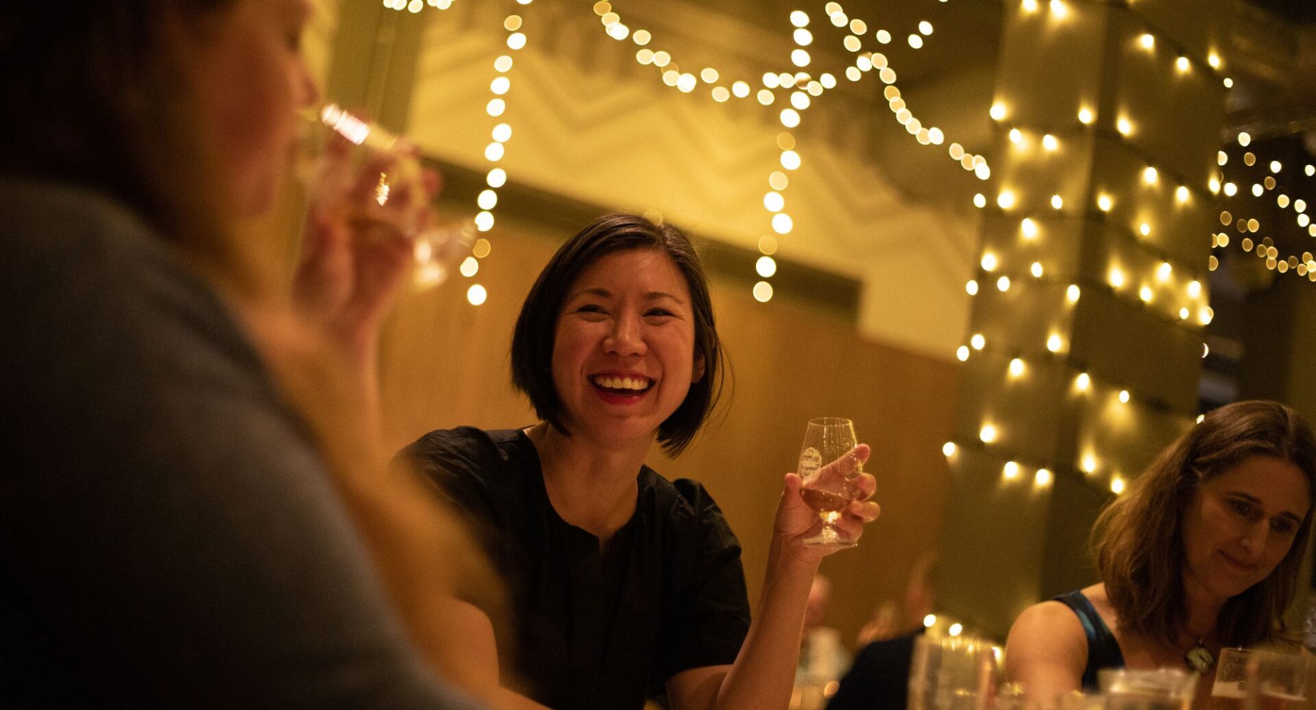 Group of people seated at a table. In the background, the walls are decorated with fairy lights.