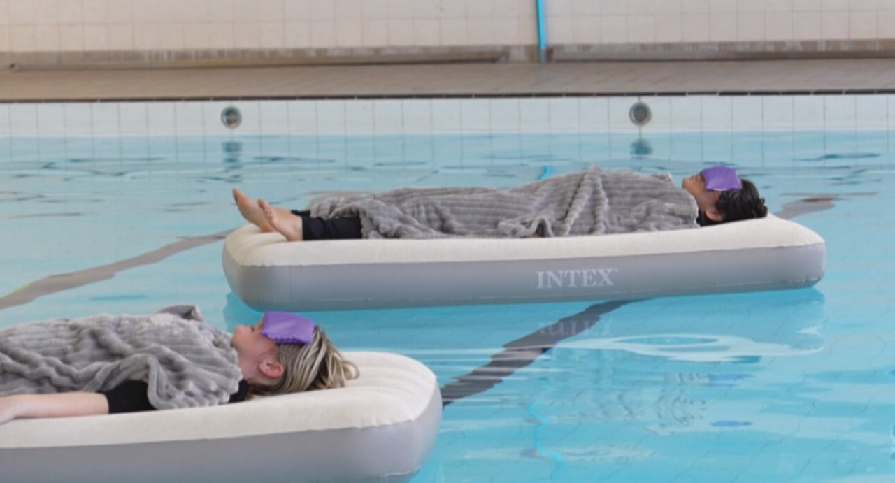 2 people covered with blankets and wearing eye masks, floating on a floating bed in a pool.