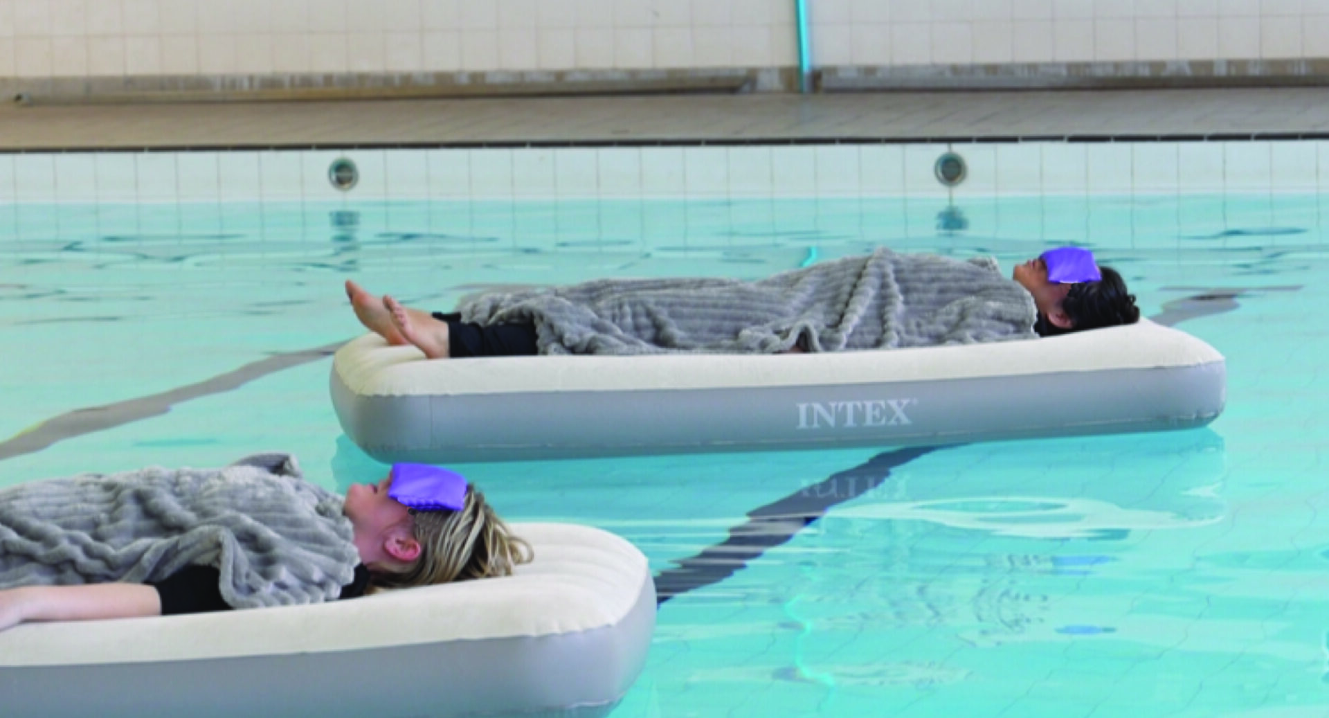2 people covered with blankets and wearing eye masks, floating on a floating bed in a pool.