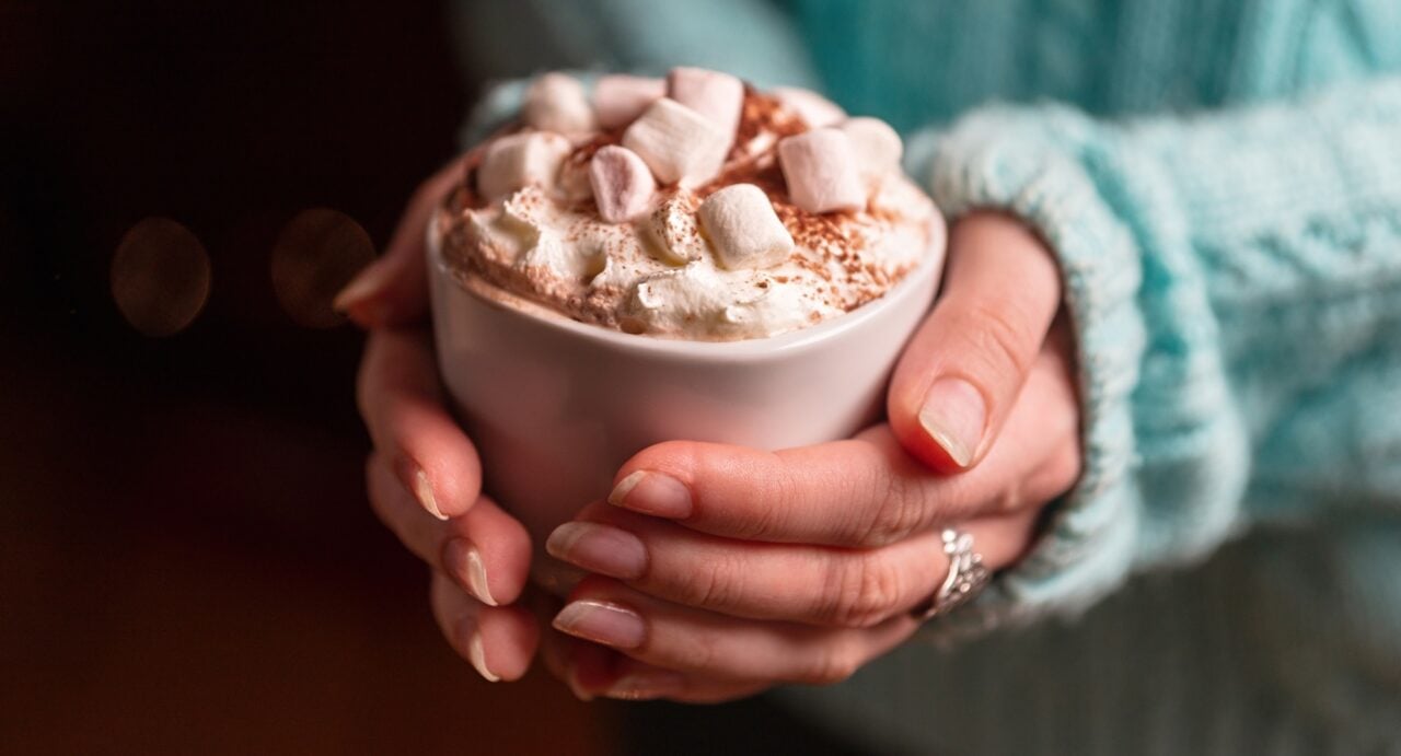 Female hands holding white ceramic mug of hot chocolate with marshmallows in hands. Green warm sweater as background.