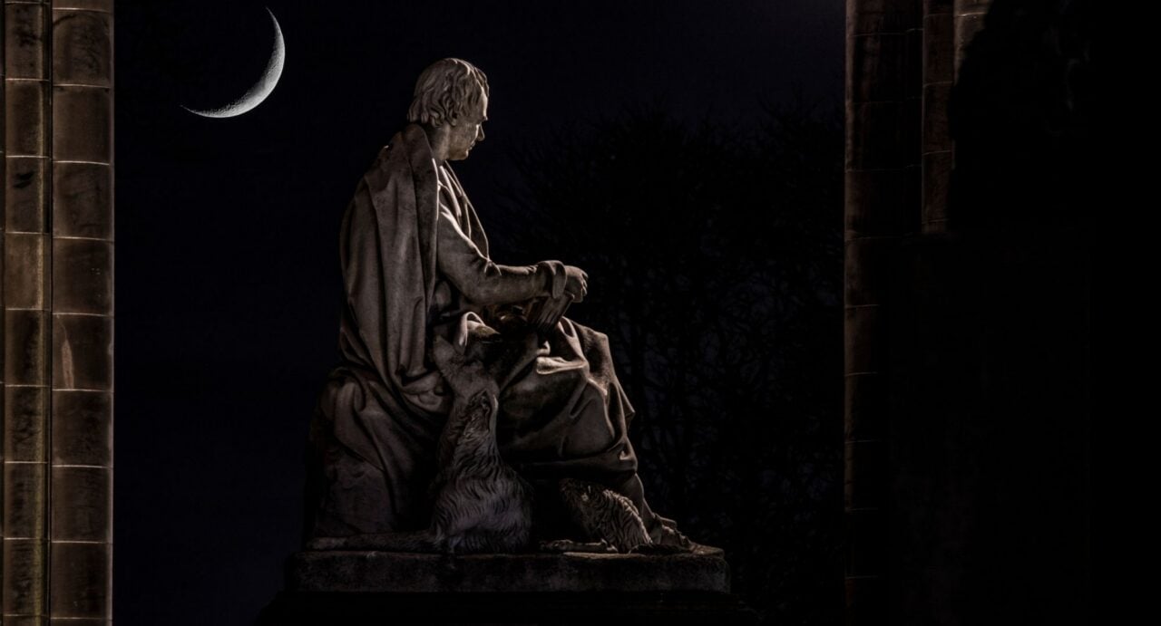 Night-time photo of a sculpture of a seated man, with a dog by his side. In the background is the silver crescent of a moon.