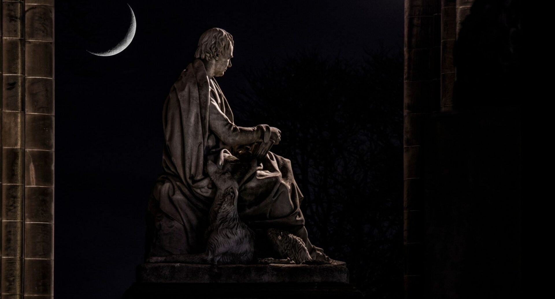 Night-time photo of a sculpture of a seated man, with a dog by his side. In the background is the silver crescent of a moon.