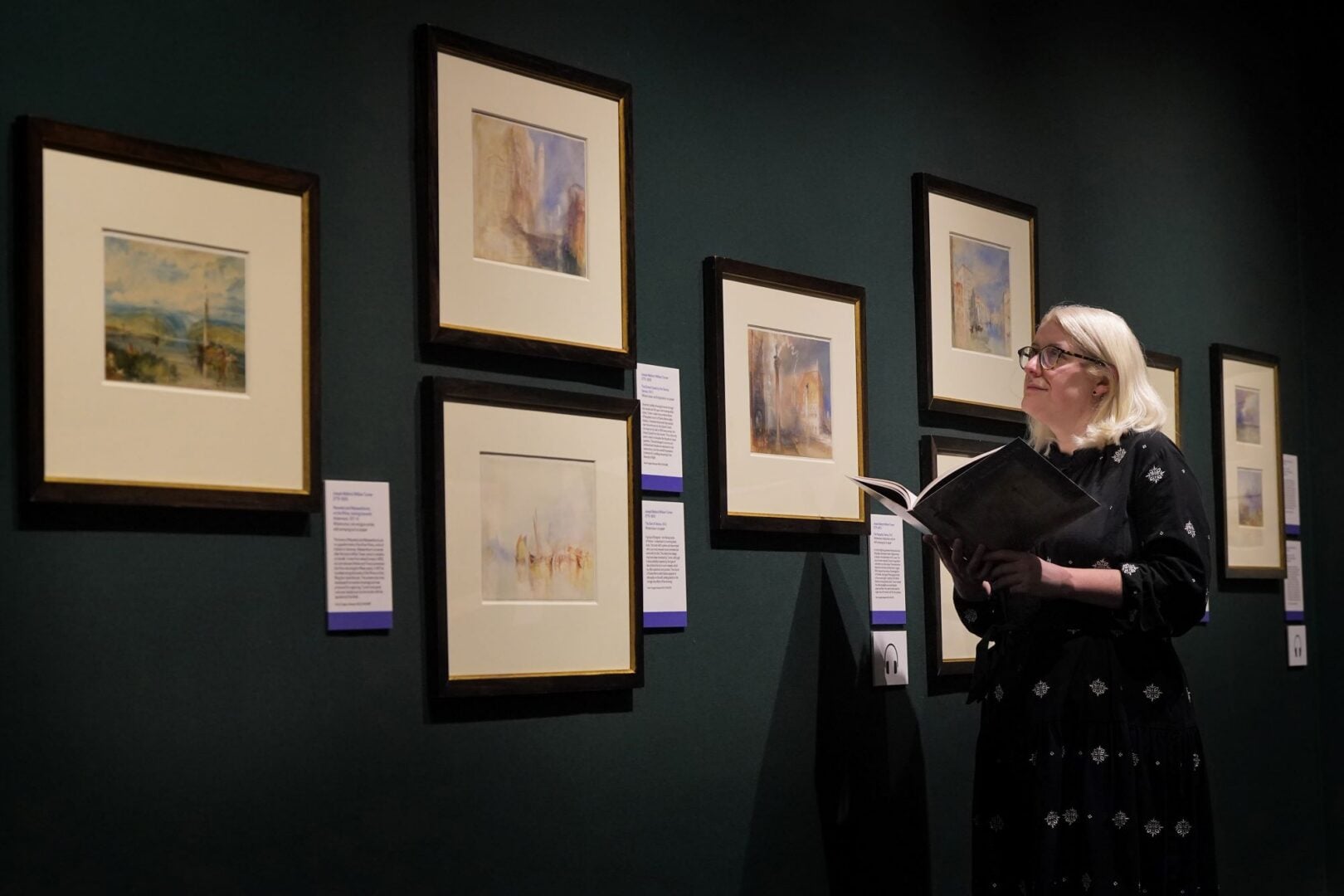 A woman holding a book and looking at paintings on a wall.