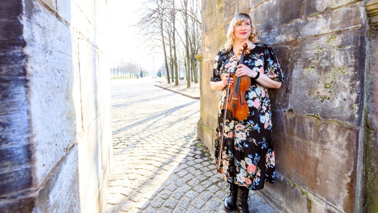Woman in long flowery dress, holding a violin, and standing leaning against a stone wall.