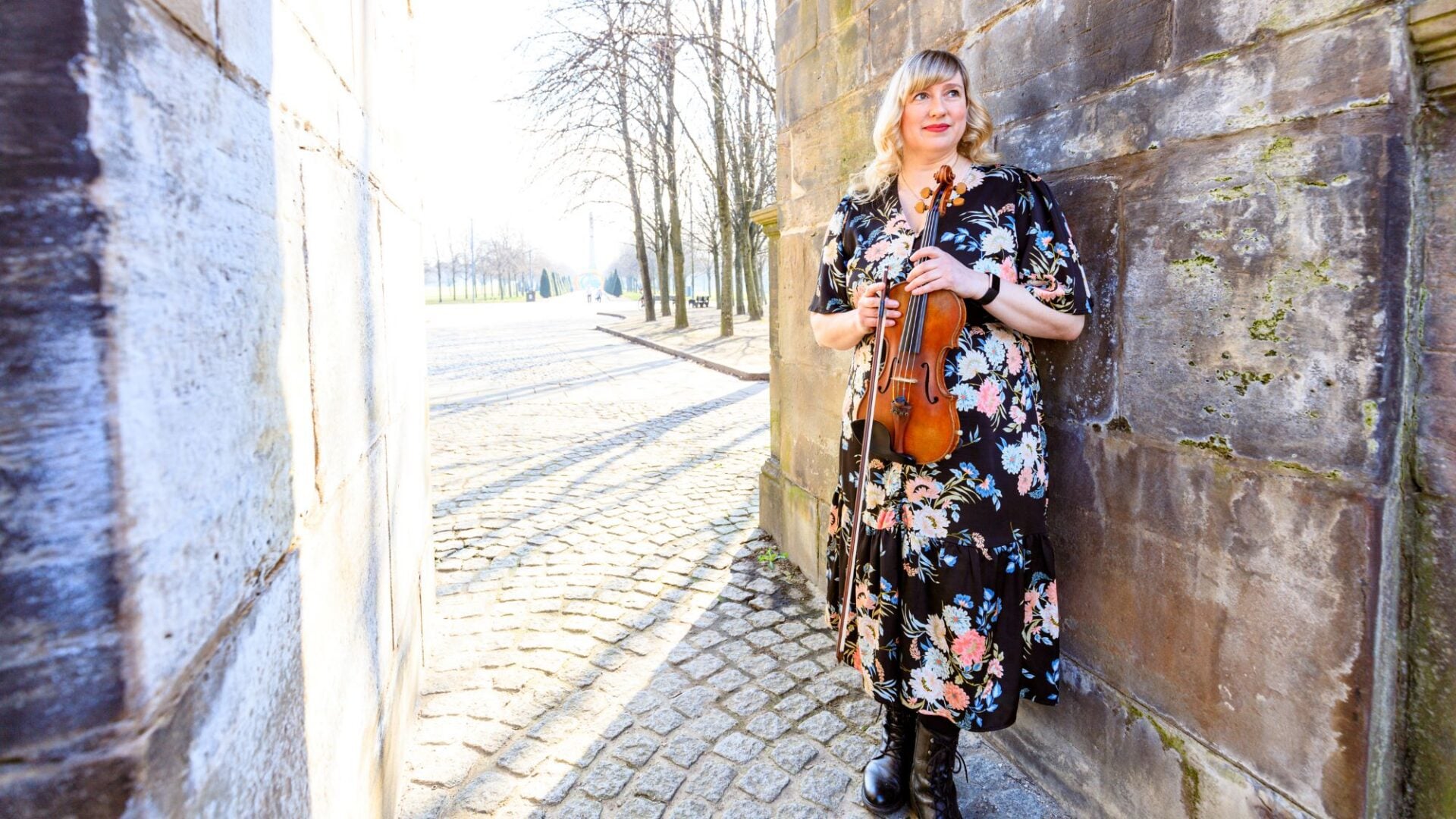 Woman in long flowery dress, holding a violin, and standing leaning against a stone wall.