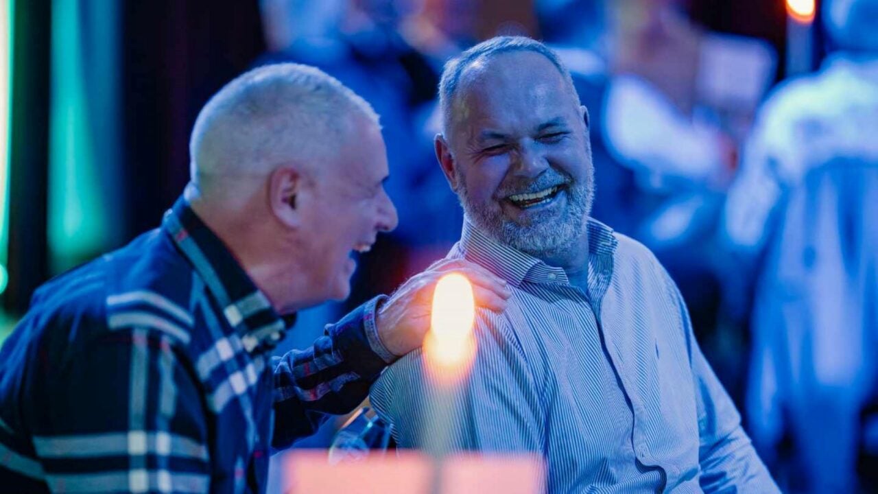 2 seated men, laughing. In front of them is a lit candle.