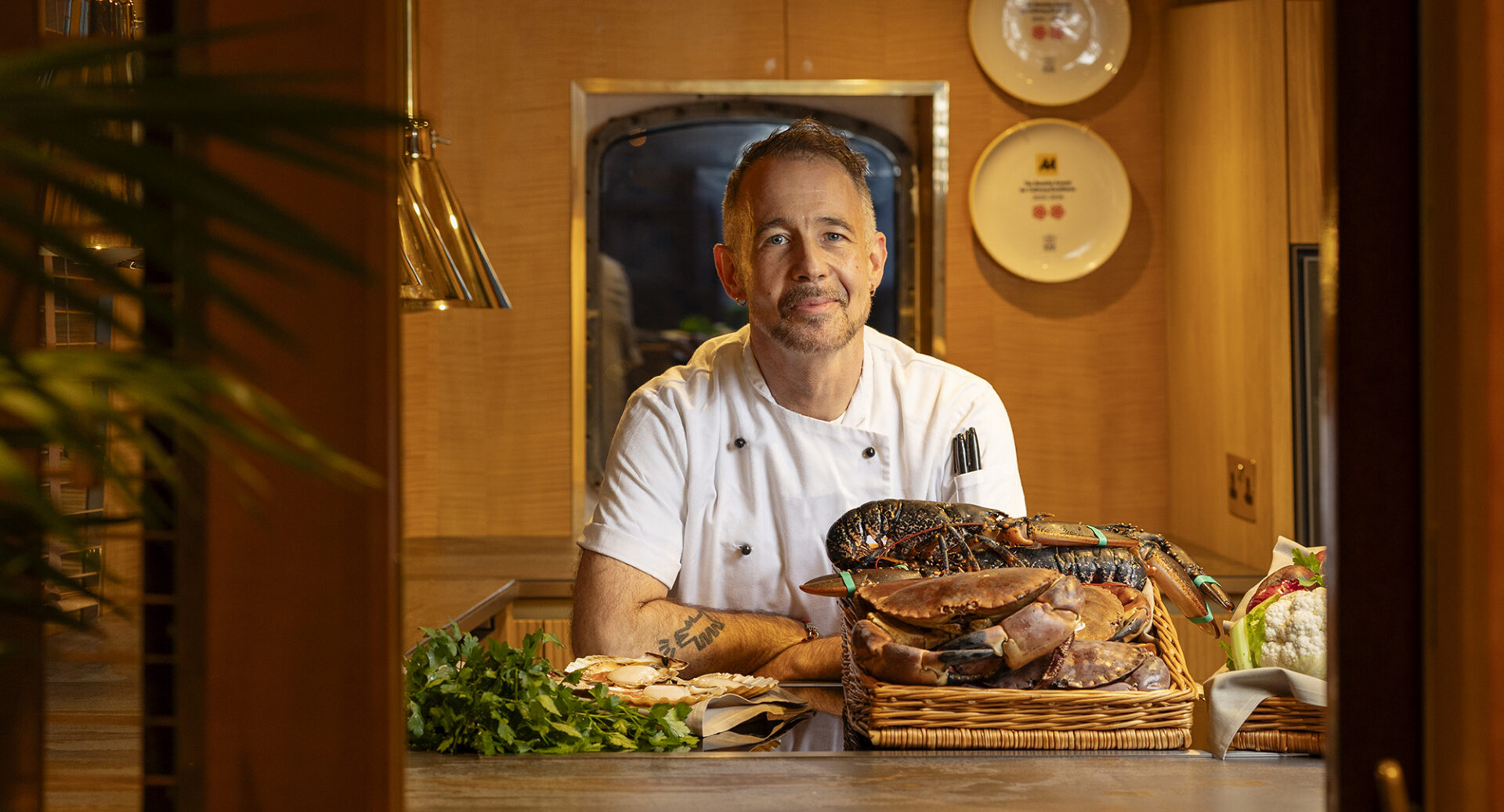 Man in chef's uniform standing in front of a basket of fresh seafood and vegetables.
