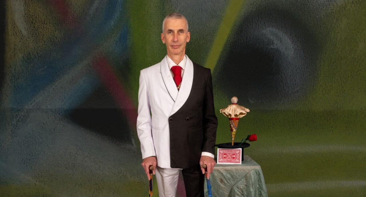 A man wearing a black and white suit, standing beside a small table.