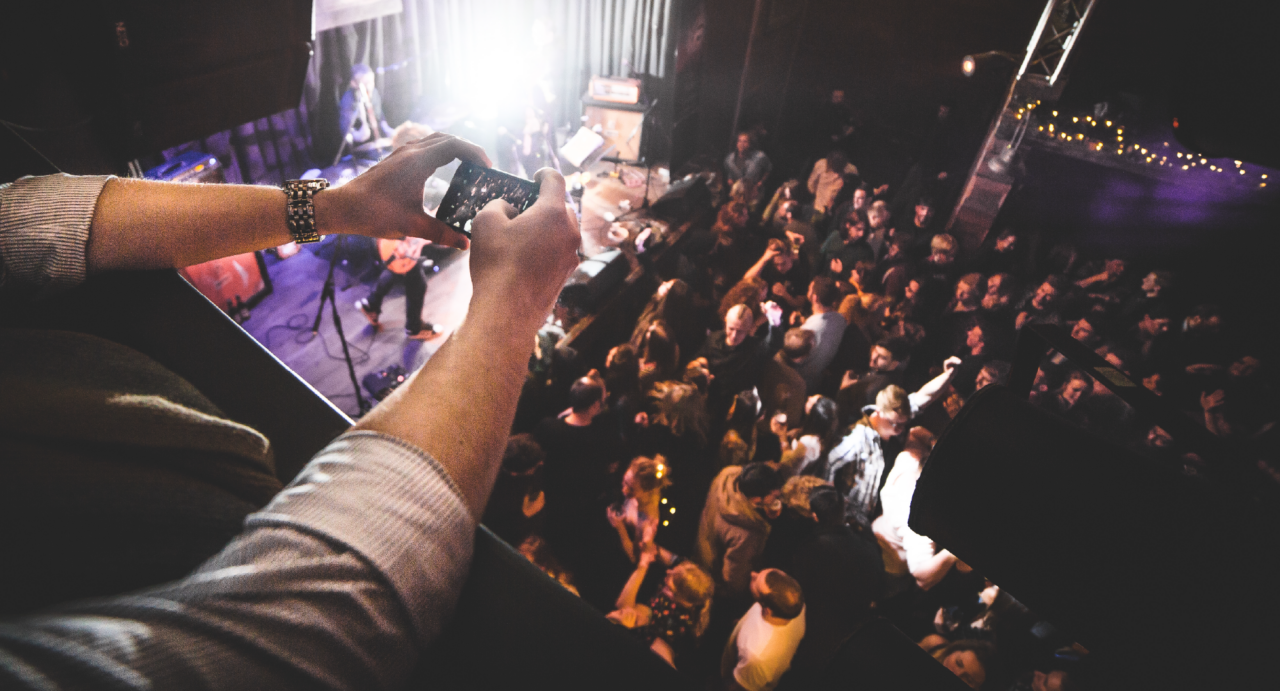 Person looking over a balcony, onto stage and floor below, which is full of people.