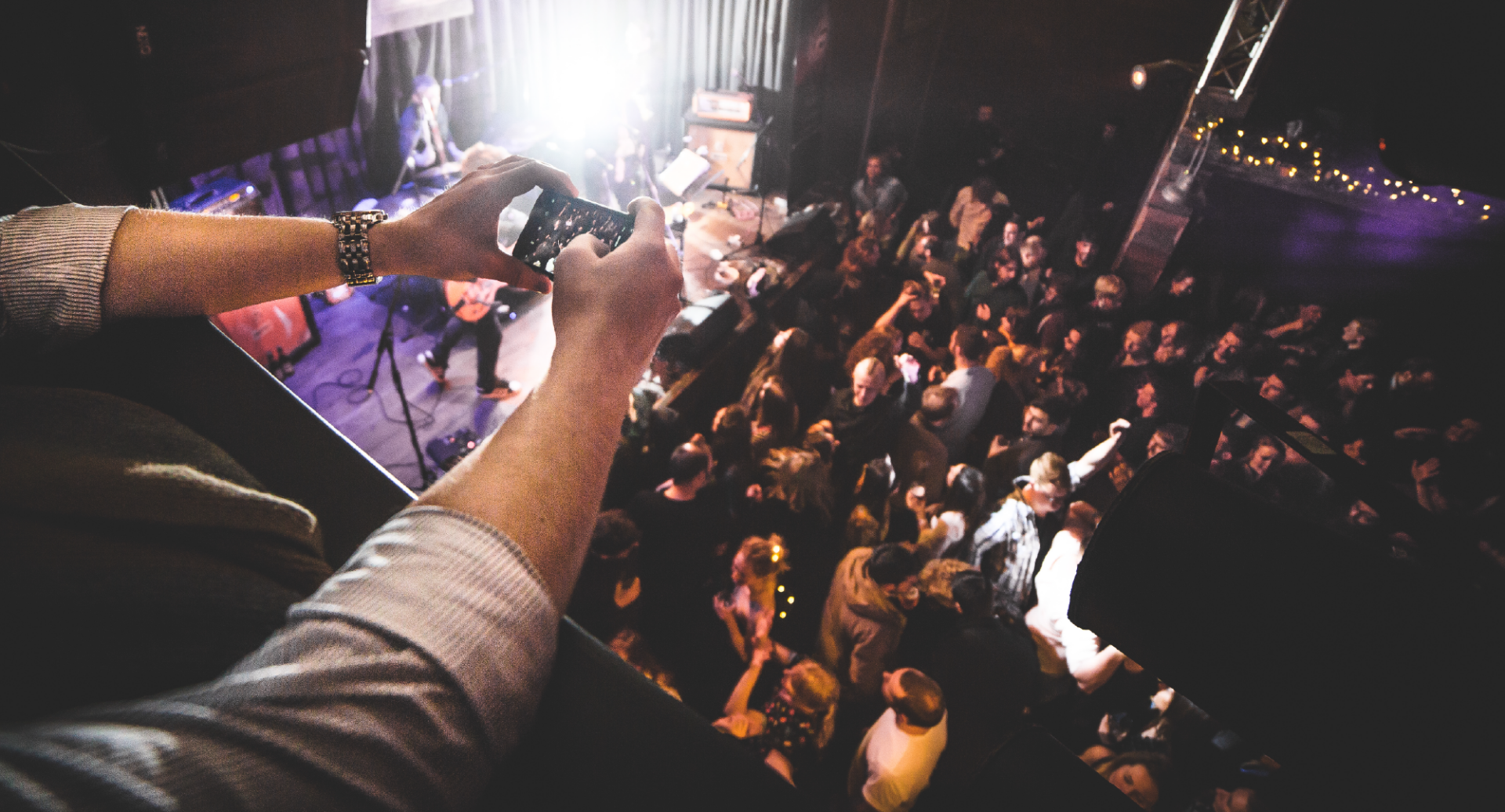 Person looking over a balcony, onto stage and floor below, which is full of people.