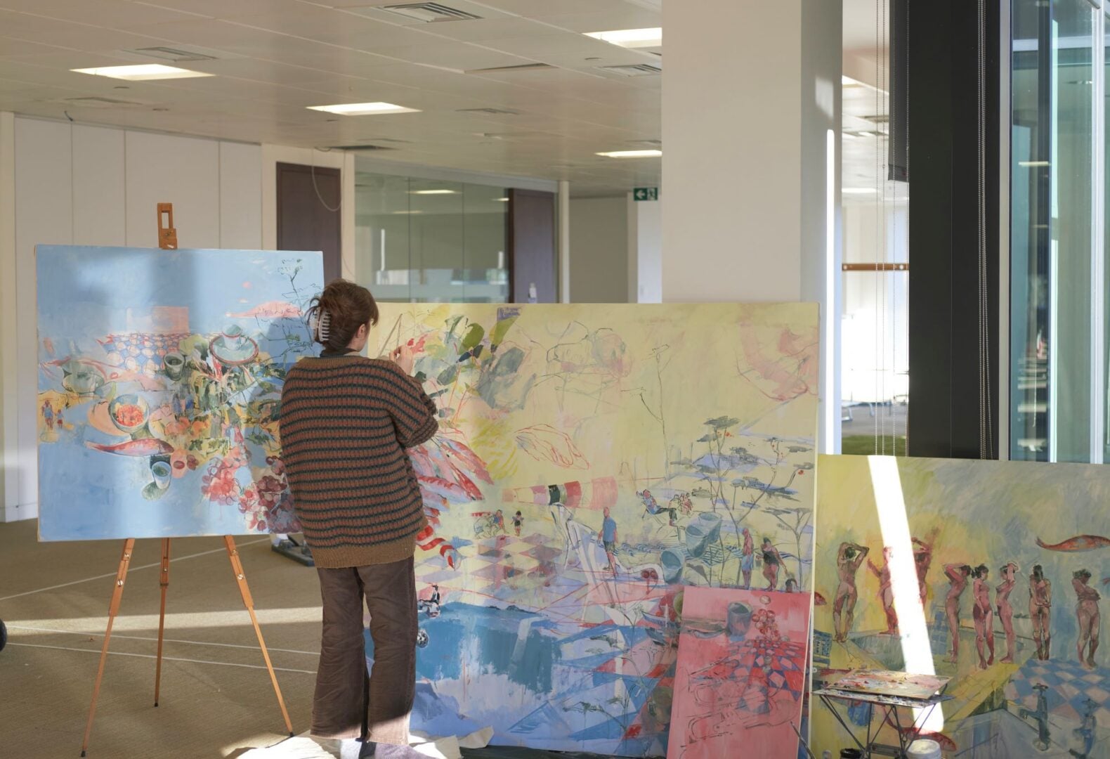 Person standing with back to camera, in an artist's studio, surrounded by painted canvases.