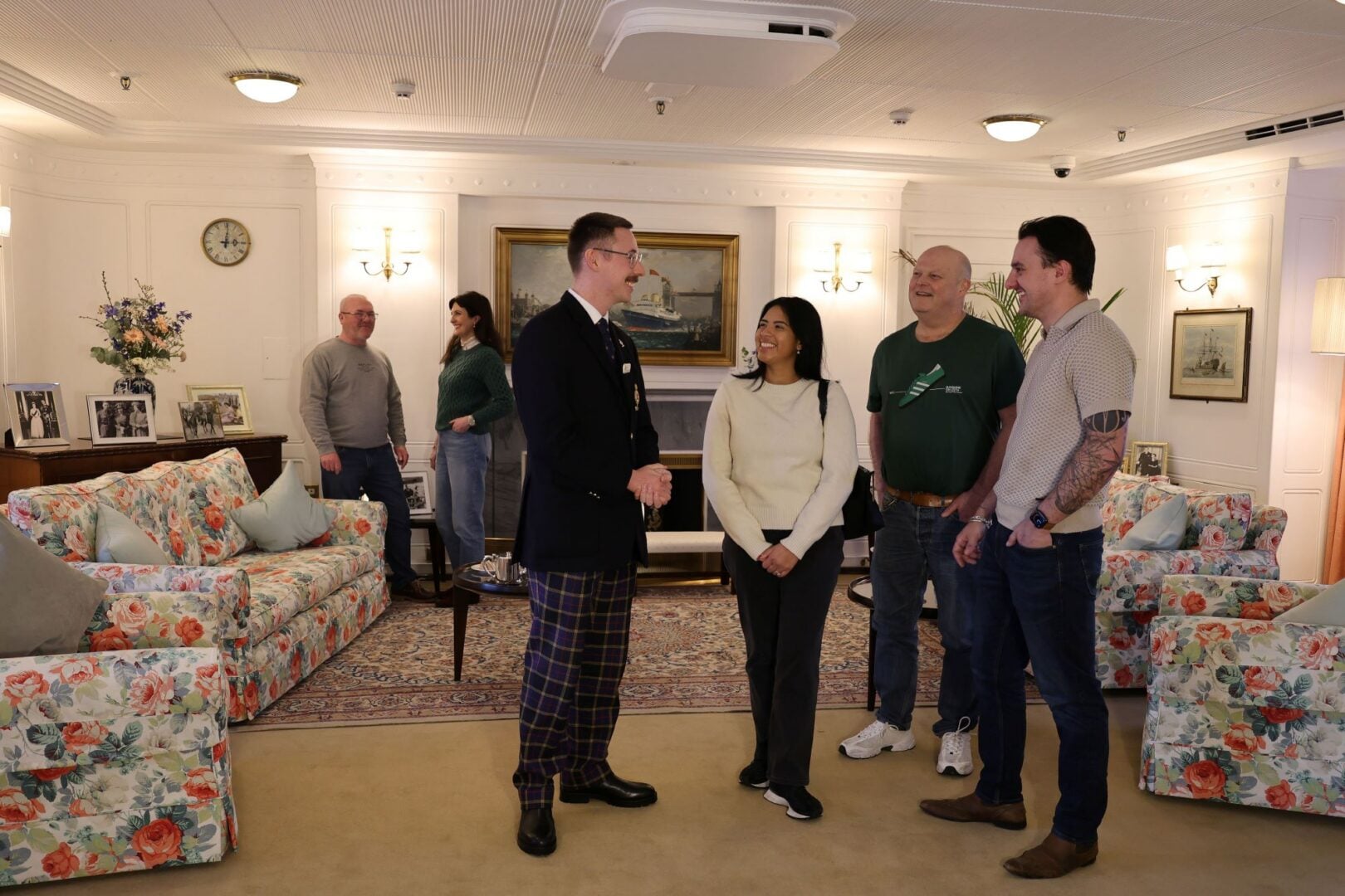 Group of people in a lounge area, complete with floral-covered couches and photos on the walls.