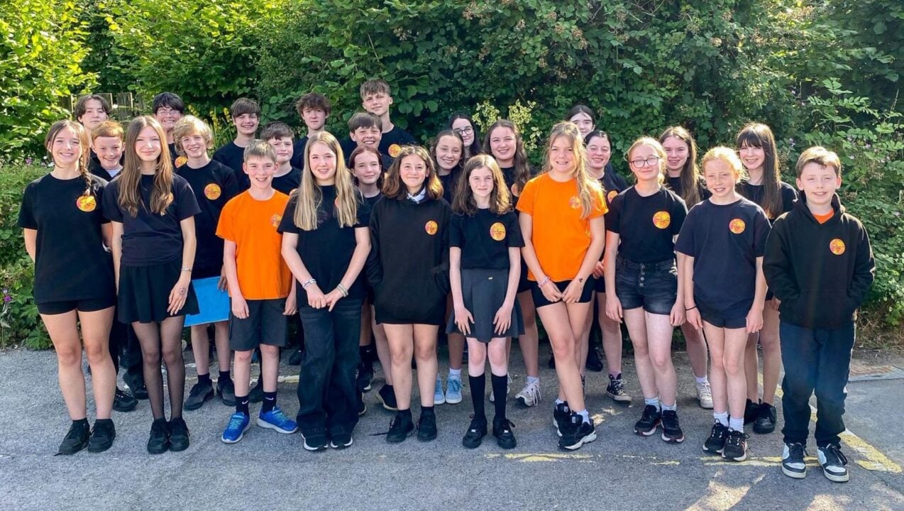 Group of young children, standing against a large green hedge.