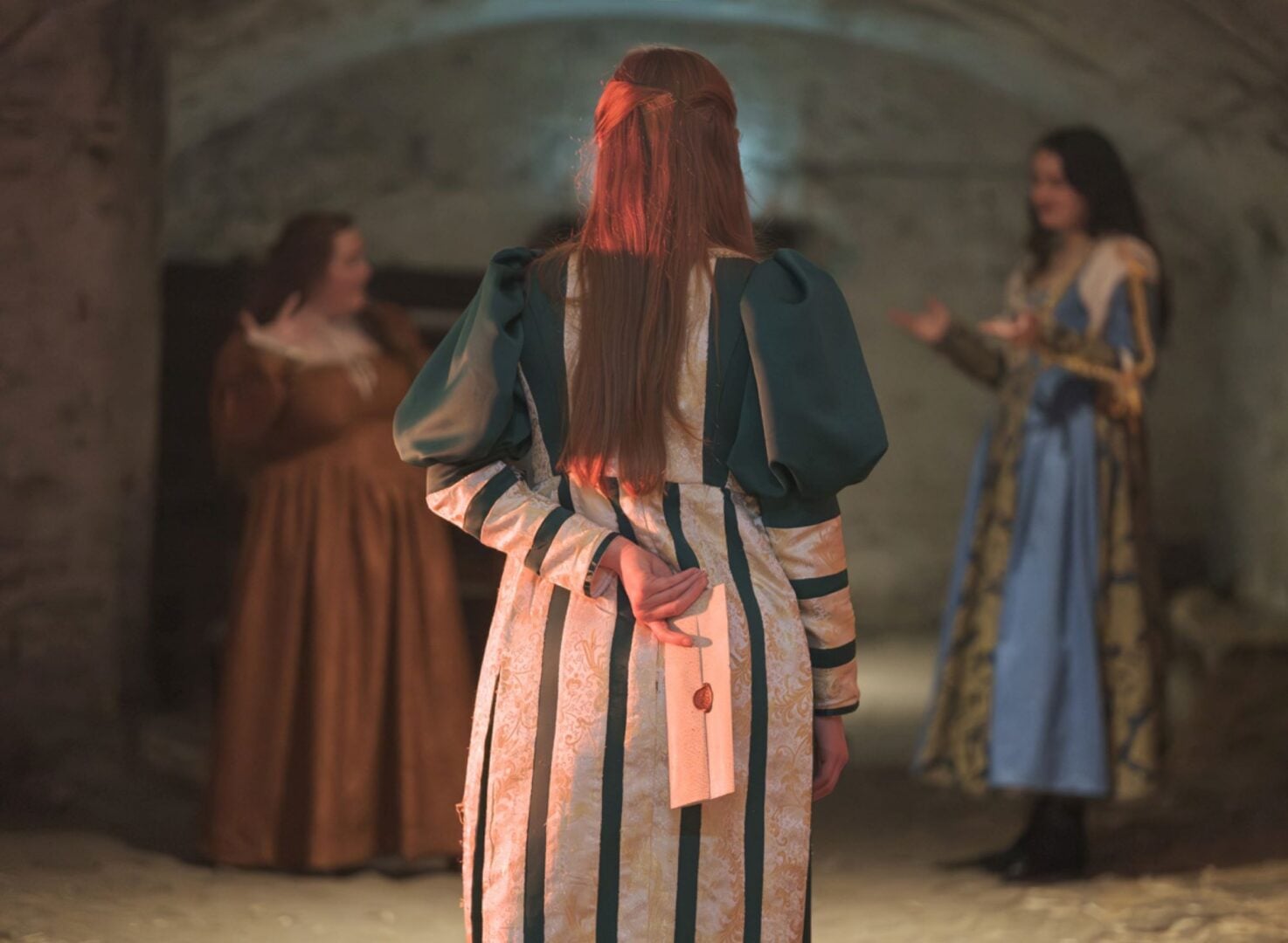 3 women, in old fashioned dress, in an underground space. One of them is holding a sealed envelope behind her back.