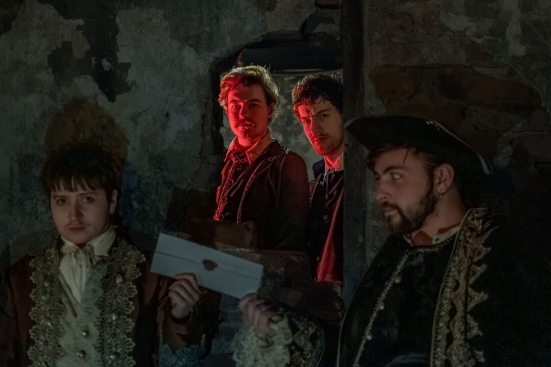 4 men in old fashioned dress, standing in an underground space. One of them is passing a sealed envelope to another.