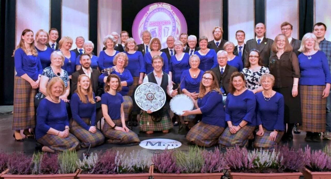 A group of people, standing and seated on stage. The 2 people in front are holding 2 large silver plates.