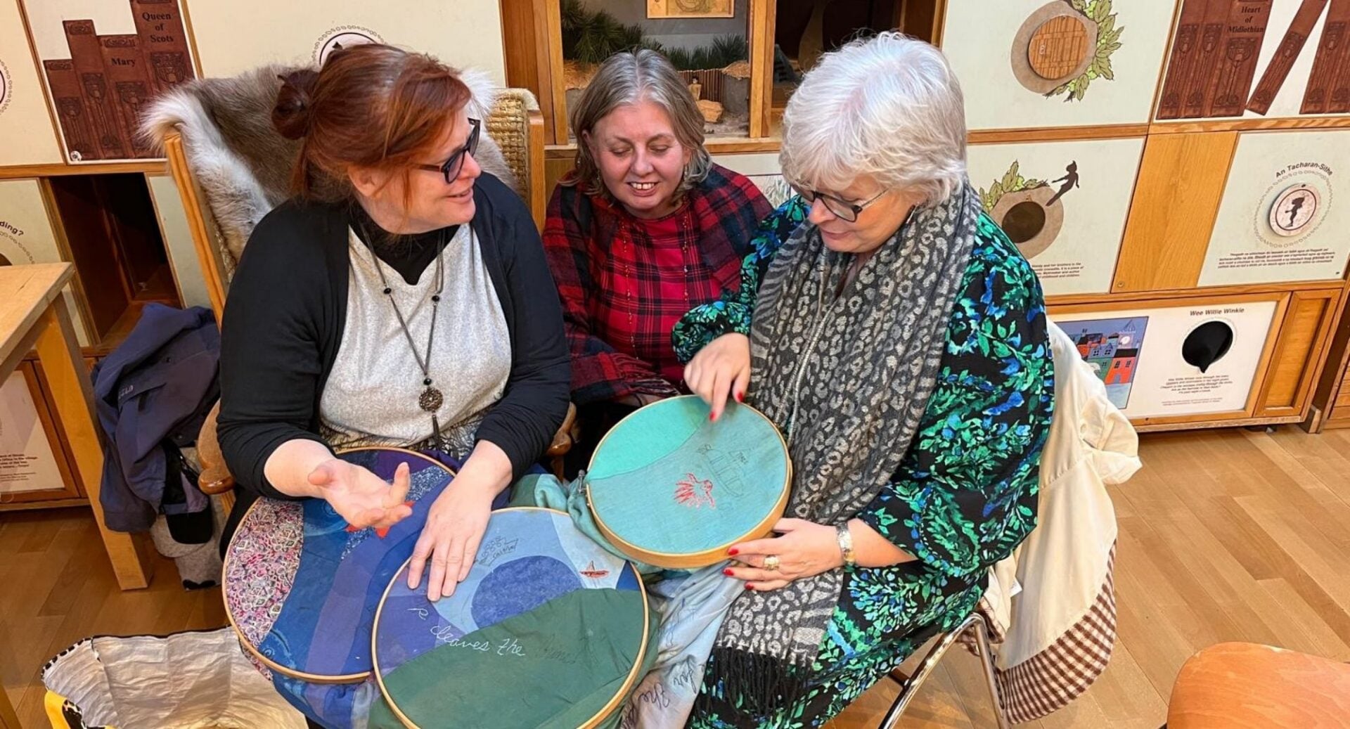 3 seated women, holding sewing materials.