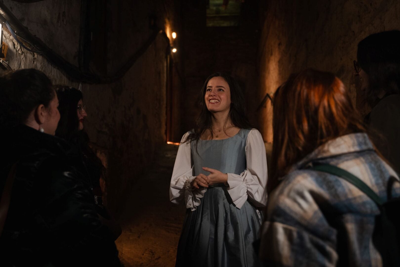 A woman wearing an old fashioned white and blue dress, in an underground setting, talking to a group of people.
