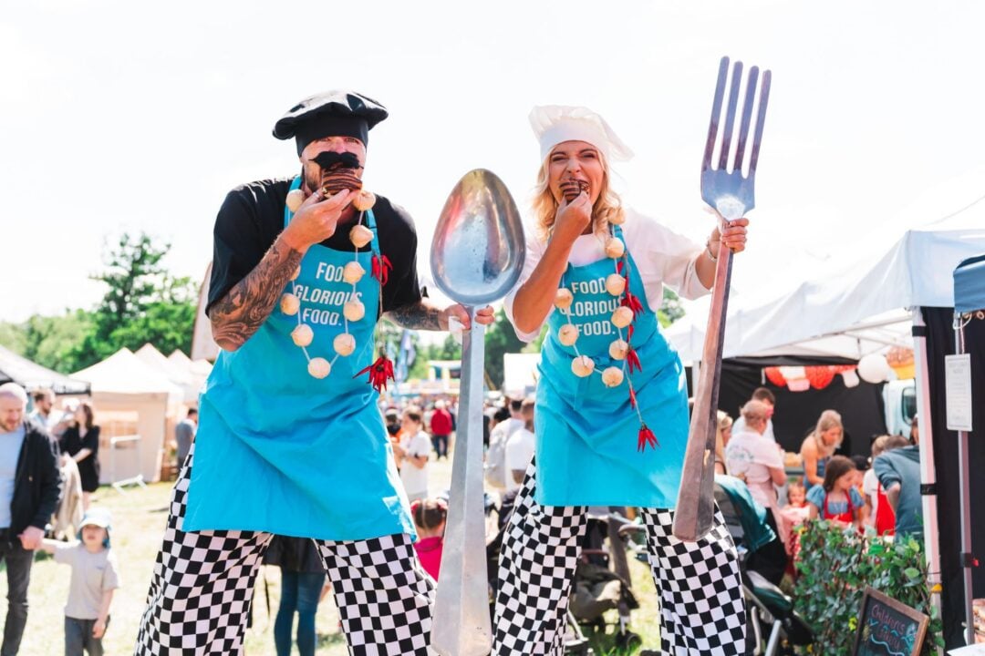 2 people dressed as chefs, holding over-sized spoons and forks.