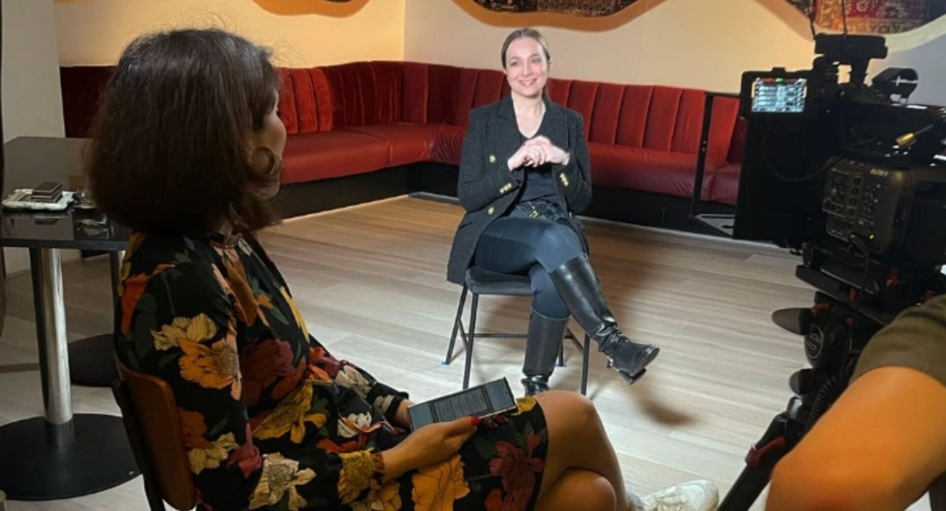 A seated woman interviewing another woman, whose back is to the camera. In the foreground is a video camera.
