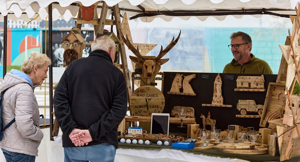 2 people looking at a stall filled with lots of wooden items.