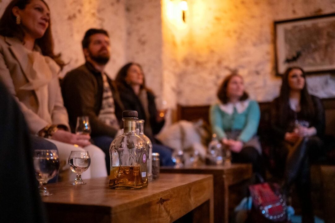 Group of people seated round small tables, on which there are bottles of whisky and glasses.