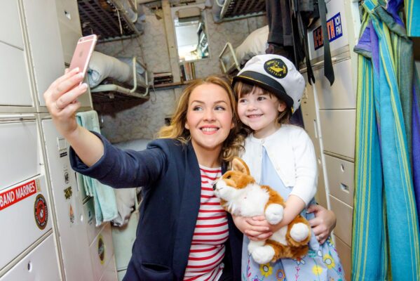 A woman and a young girl take a selfie on abord a ship. The girl is wearing a captain's hat and holding a toy corgi dog.