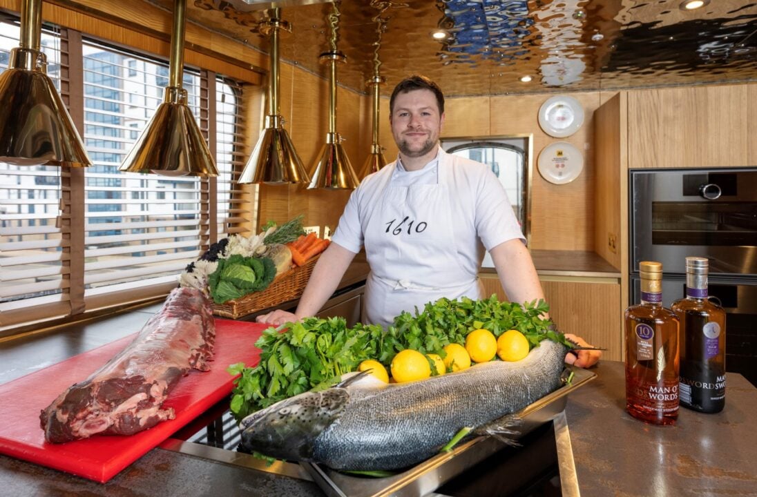 A man in a chef uniform, standing in a kitchen. In front of him are a selection of food and drink ingredients.
