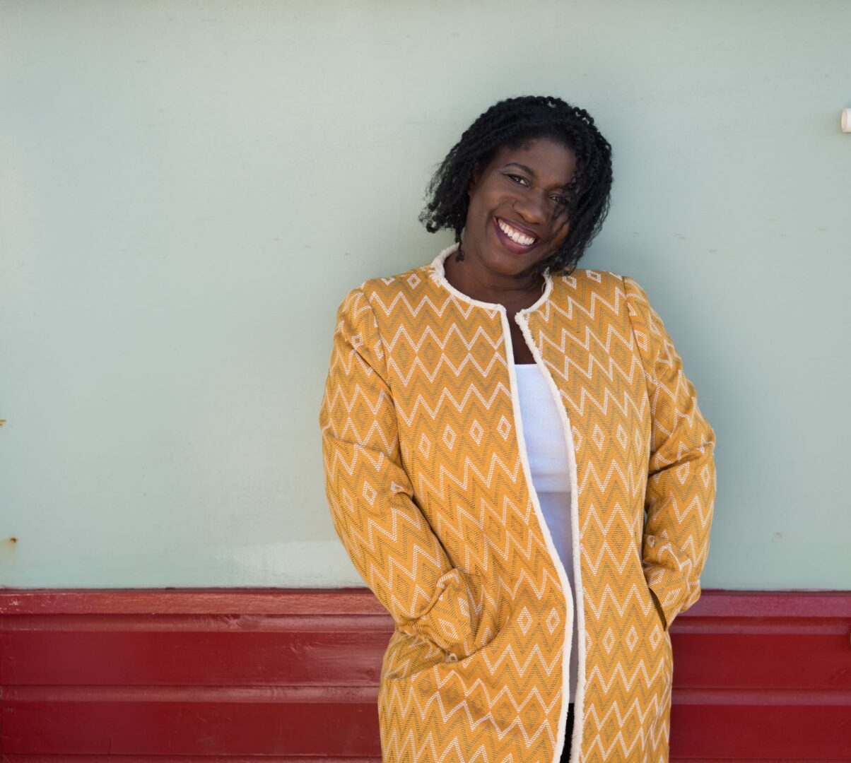 Women in orange and white long jacket leaning against a wall.