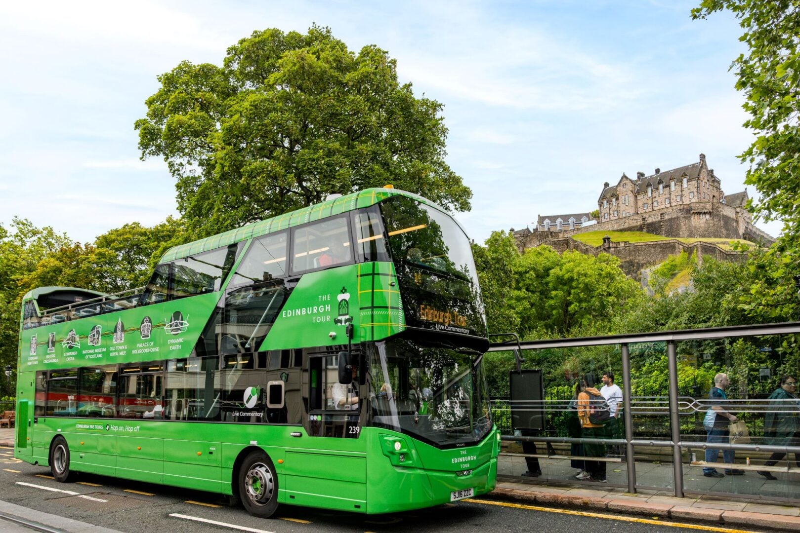 A green open-top bus, at a bus stop. In the background can be seen a castle at the top of a hill.