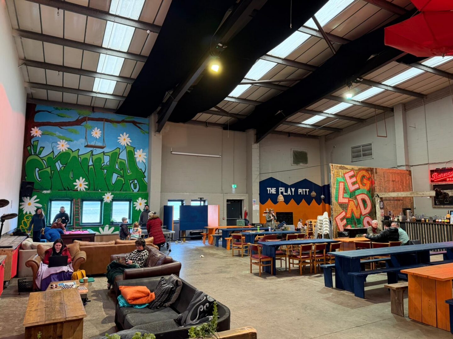 People sat around soft seats and bench tables in a warehouse-style building.
