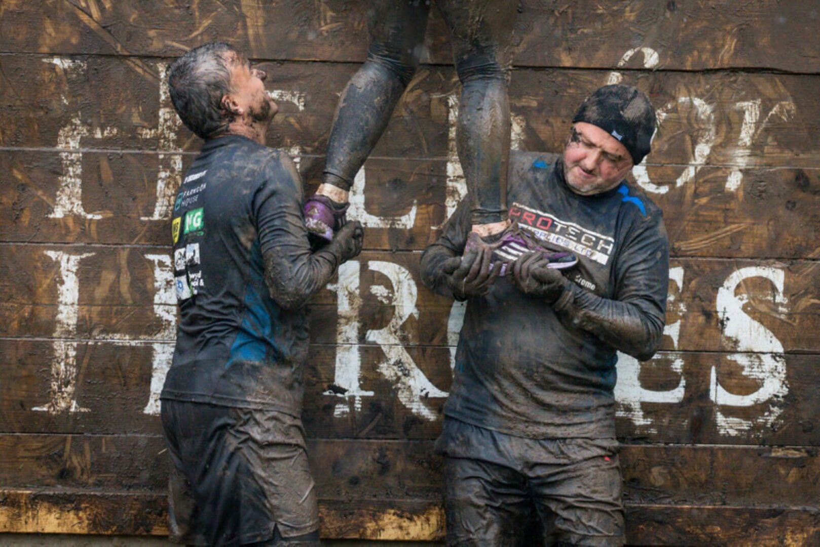 2 men in muddy clothes holding someone's legs to get over a wall.