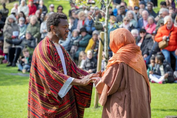 2 people standing in a grassy area, in front of a crowd of people.