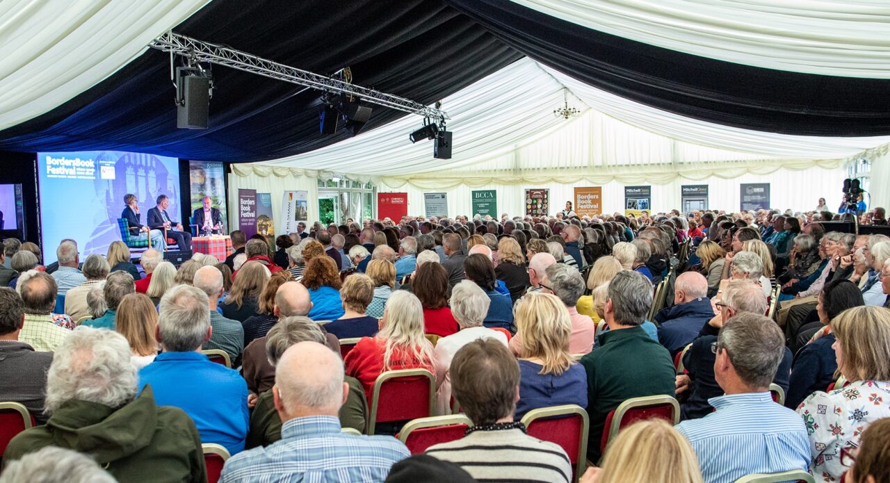 A group of seated people in a marque watching 3 people on a stage.