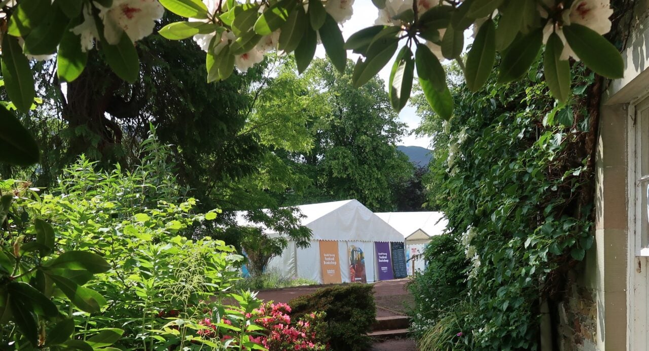 A white marque seen through a gap in green hedges and bushes.