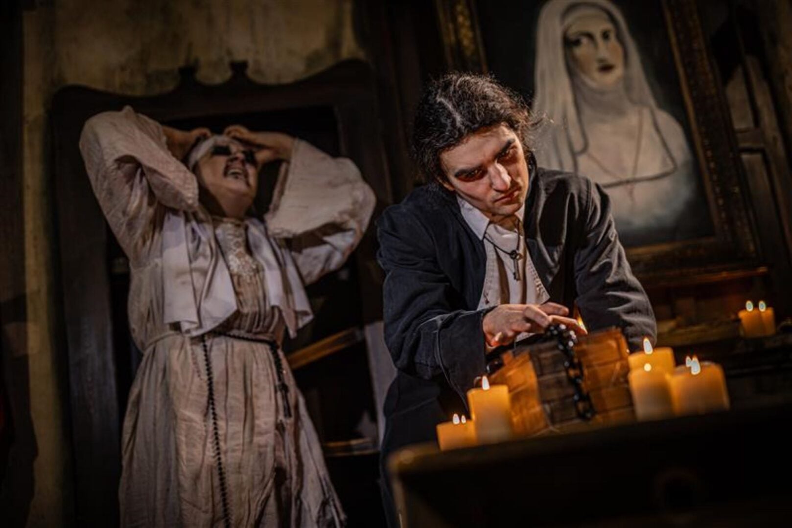 A brooding man sitting in front of lit candles, and a nun holding her head in the background.