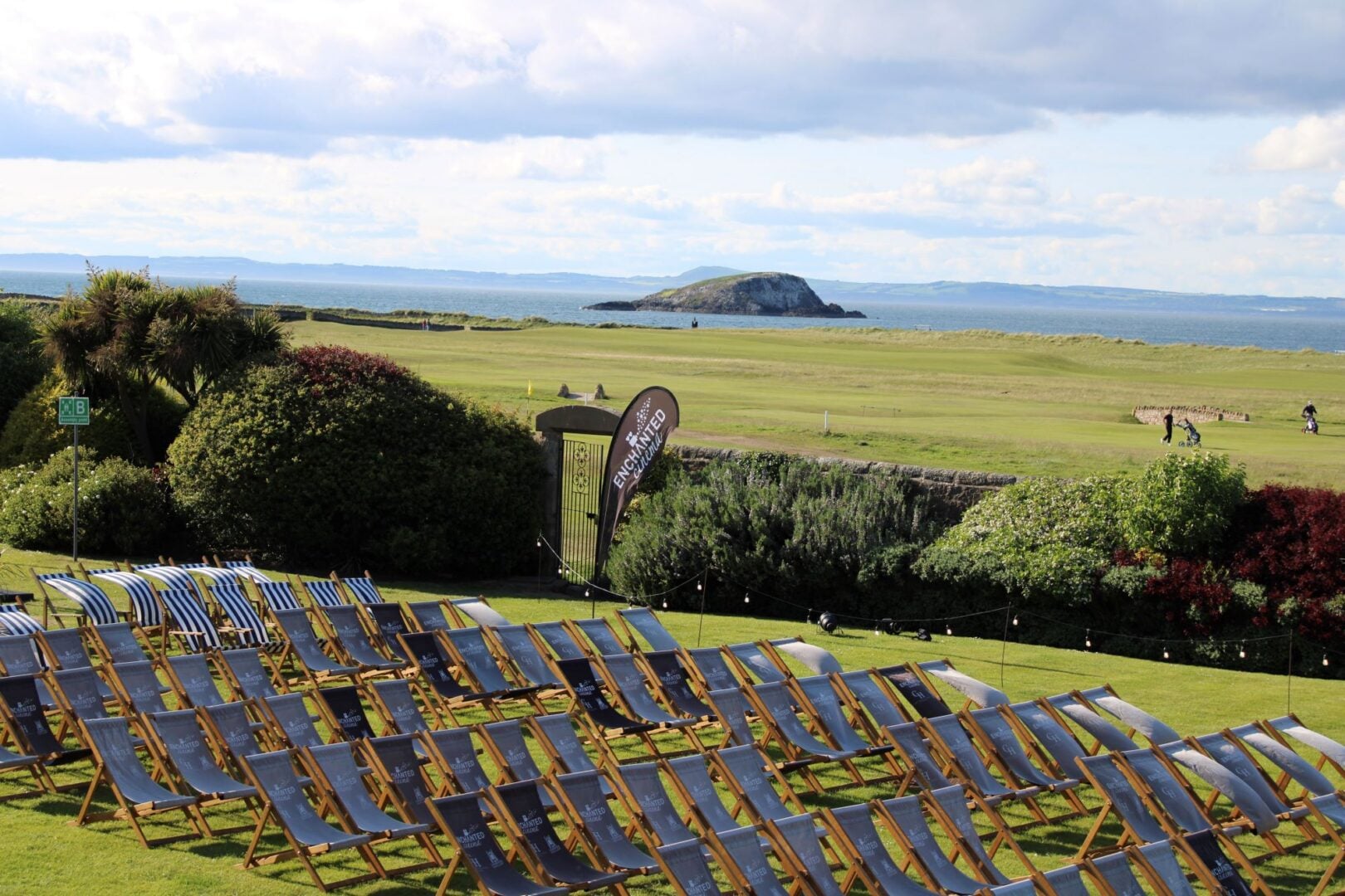 Rows of deckchairs set up on a grassy field, overlooking a golf course and the coast.