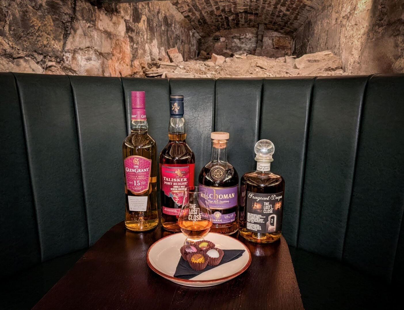 4 bottles of whisky sitting on a table, alongside a glass of whisky and 4 chocolates.