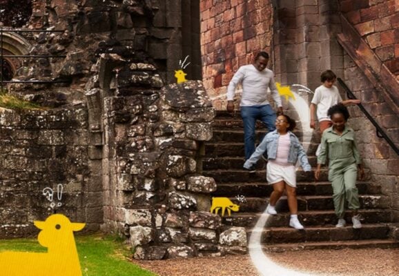 2 adults and 2 children running down the steps of a stone building, surrounded by cartoon lambs.
