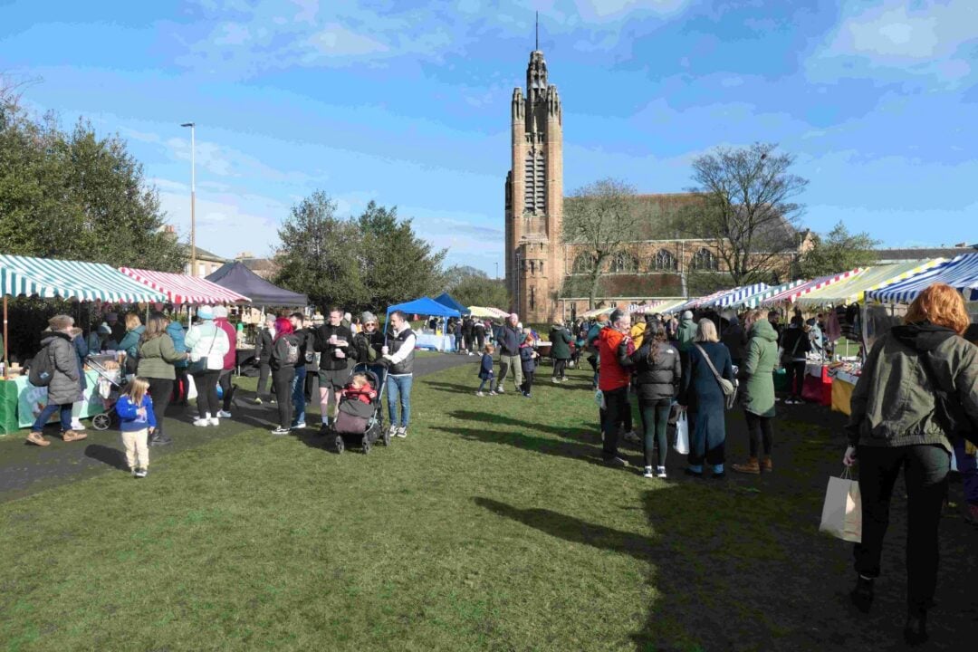 People browsing stalls at an outdoor market. In the distance is a church with a tall steeple.