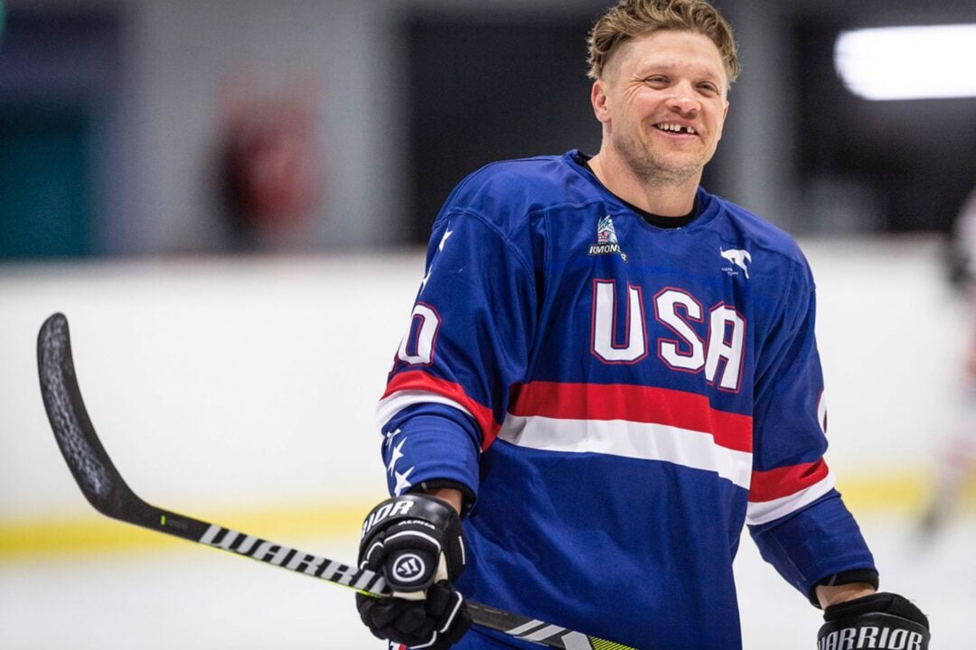 A man wearing a padded red, blue and white ice hockey top, and holding an ice hockey stick.