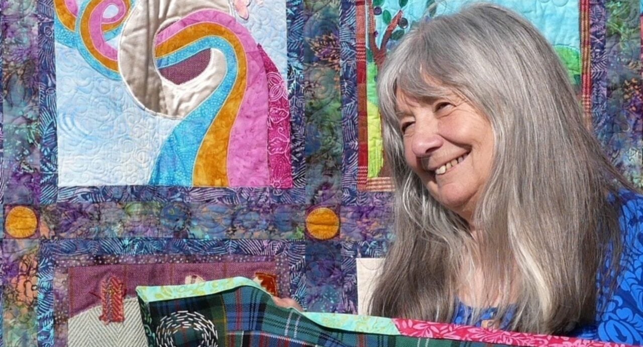 A woman with long grey hair beside a multi-coloured quilt.