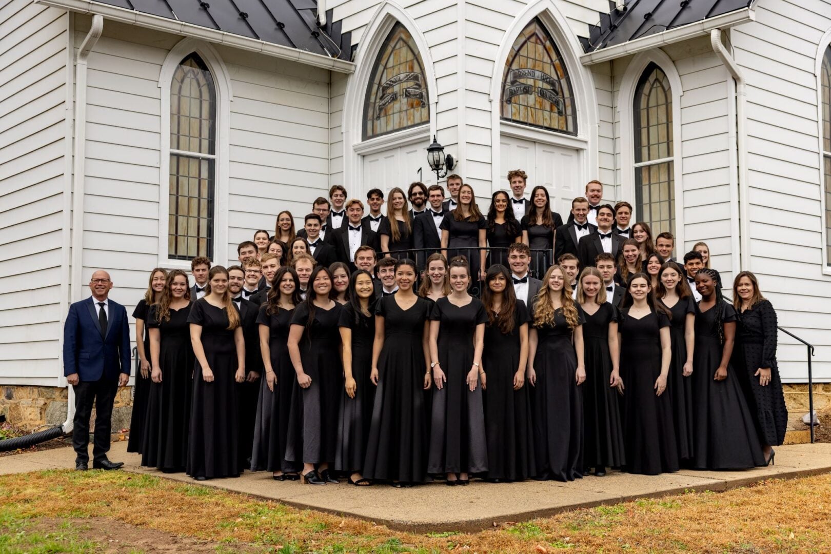 A group of people, all wearing black, standing outside a white building, with stained glass windows.