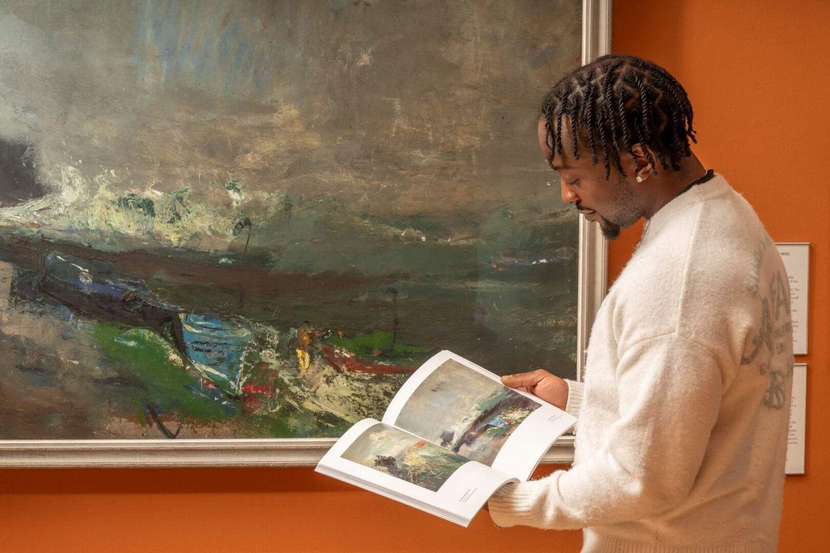 A man looking at a painting in a book, while standing beside a wall, on which the original painting can be seen.